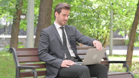 Téléchargez les photos : Jeune homme d'affaires à venir, assis sur le banc et travaillant sur ordinateur portable - en image libre de droit