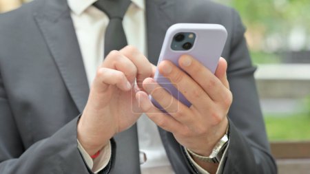 Téléchargez les photos : Gros plan sur un jeune homme d'affaires naviguant sur un téléphone intelligent assis à l'extérieur sur un banc - en image libre de droit