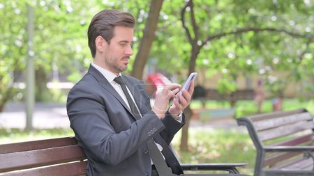 Photo for Young Businessman Browsing Internet Smartphone while Sitting Outdoor on a Bench - Royalty Free Image