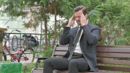 Téléchargez les photos : Moyen Age Homme d'affaires assis en plein air avec maux de tête - en image libre de droit