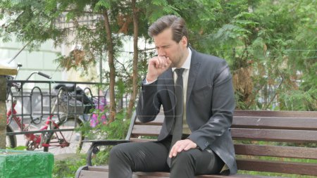 Téléchargez les photos : Toux moyen âge homme d'affaires assis en plein air sur banc - en image libre de droit