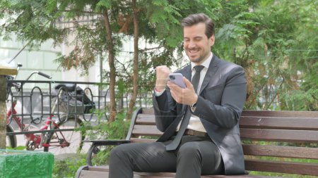 Foto de Empresario de mediana edad celebrando el éxito en el teléfono inteligente mientras está sentado al aire libre en un banco - Imagen libre de derechos