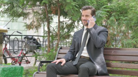 Foto de Empresario de mediana edad hablando por teléfono mientras está sentado al aire libre en un banco - Imagen libre de derechos