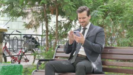 Téléchargez les photos : Moyen Age Homme d'affaires choqué par la perte sur Smartphone tout en étant assis à l'extérieur sur un banc - en image libre de droit