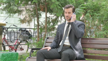 Téléchargez les photos : Bouleversé Moyen Age homme d'affaires se disputant au téléphone tout en étant assis en plein air sur un banc - en image libre de droit