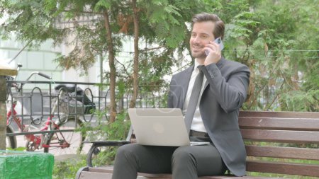 Photo for Middle Aged Businessman Talking on Phone and Using Laptop Outdoor - Royalty Free Image