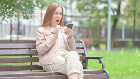 Foto de Anciana mayor sorprendida por la pérdida en el teléfono mientras está sentada al aire libre en un banco - Imagen libre de derechos
