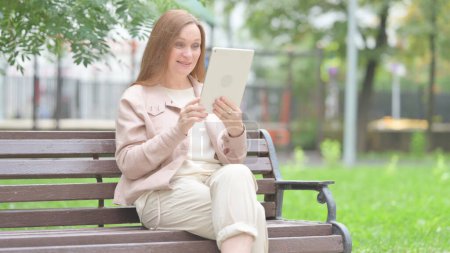 Téléchargez les photos : Vieille femme âgée faisant du chat vidéo sur tablette tout en étant assise en plein air sur un banc - en image libre de droit