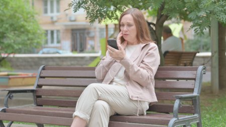 Téléchargez les photos : Vieille femme moderne face à la perte sur Smartphone tout en étant assis sur le banc - en image libre de droit
