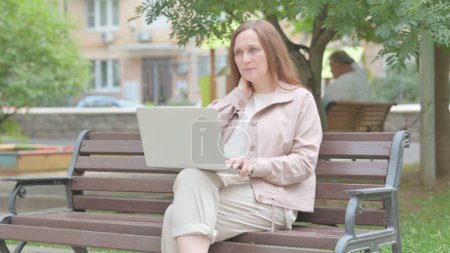 Téléchargez les photos : Vieille femme moderne avec douleur au cou en utilisant un ordinateur portable en plein air - en image libre de droit