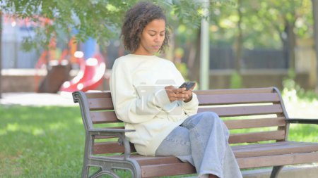 Photo for Young African Woman using Phone while Sitting Outdoor on a Bench - Royalty Free Image