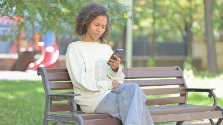 Photo for Young African Woman Shocked by Loss on Phone while Sitting Outdoor on a Bench - Royalty Free Image