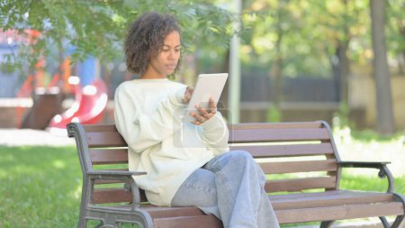 Téléchargez les photos : Jeune femme africaine en utilisant une tablette tout en étant assis en plein air sur un banc - en image libre de droit