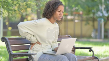 Téléchargez les photos : Jeune femme africaine utilisant un ordinateur portable avec douleur au dos tout en étant assis à l'extérieur - en image libre de droit