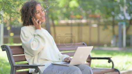Téléchargez les photos : Jeune femme africaine parlant au téléphone et utilisant un ordinateur portable en plein air - en image libre de droit