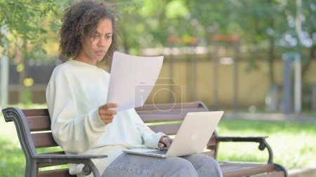 Téléchargez les photos : Jeune femme africaine travaillant sur des documents et ordinateur portable en plein air - en image libre de droit
