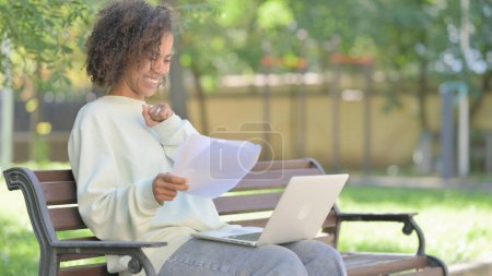 Téléchargez les photos : Jeune femme africaine célébrant tout en travaillant sur ordinateur portable et documents en plein air - en image libre de droit