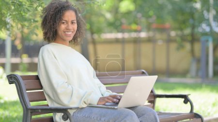 Téléchargez les photos : Jeune femme africaine souriant à la caméra tout en travaillant sur ordinateur portable en plein air - en image libre de droit