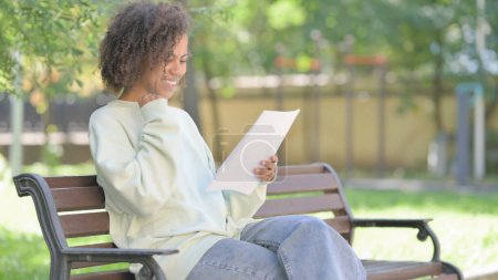 Téléchargez les photos : Jeune femme africaine célébrant et lisant des documents tout en étant assise en plein air - en image libre de droit