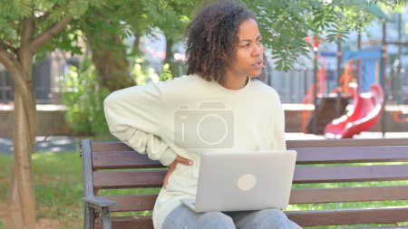 Téléchargez les photos : Femme africaine occasionnelle utilisant un ordinateur portable avec douleur au dos en plein air - en image libre de droit