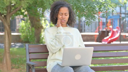 Téléchargez les photos : Femme africaine occasionnelle avec mal de dents travaillant sur ordinateur portable en plein air - en image libre de droit