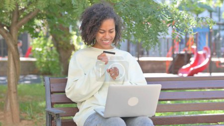 Téléchargez les photos : Femme africaine occasionnelle excitée pour réussir ses achats en ligne sur ordinateur portable en plein air - en image libre de droit