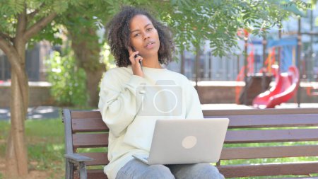 Téléchargez les photos : Femme africaine occasionnelle parlant au téléphone et utilisant un ordinateur portable en plein air - en image libre de droit