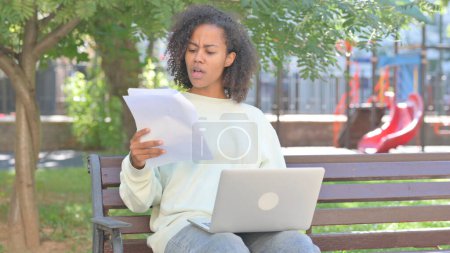 Téléchargez les photos : Casual African Woman se sentant bouleversé tout en lisant contrat et en utilisant ordinateur portable en plein air - en image libre de droit