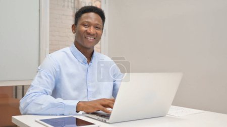 Foto de Empresario africano sonriendo a la cámara mientras trabaja en el ordenador portátil en la oficina - Imagen libre de derechos
