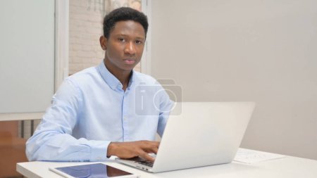 Téléchargez les photos : Homme d'affaires africain regardant la caméra tout en travaillant sur ordinateur portable dans le bureau - en image libre de droit