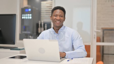 Téléchargez les photos : Africain homme d'affaires souriant à la caméra tout en travaillant sur ordinateur portable dans le bureau - en image libre de droit