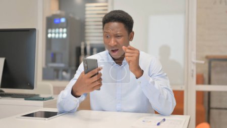Photo for African Businessman Shocked by Loss on Phone in Office - Royalty Free Image