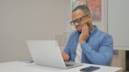 Téléchargez les photos : Homme d'affaires africain avec mal de dents Travailler sur ordinateur portable dans Office - en image libre de droit