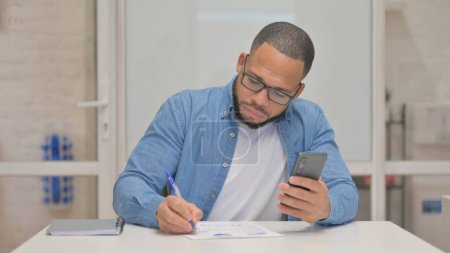 Téléchargez les photos : Homme de race mixte travaillant sur des documents avec Smartphone - en image libre de droit