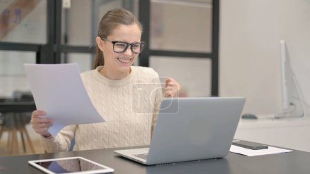 Téléchargez les photos : Jeune femme célébrant tout en utilisant un ordinateur portable et des documents - en image libre de droit