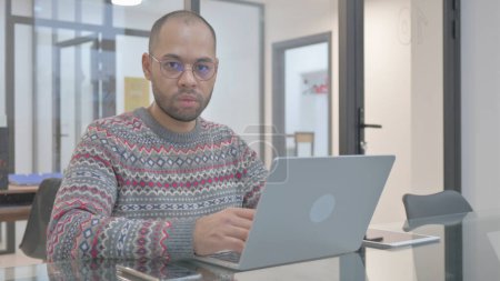 Téléchargez les photos : Jeune homme hispanique regardant la caméra tout en travaillant sur ordinateur portable - en image libre de droit