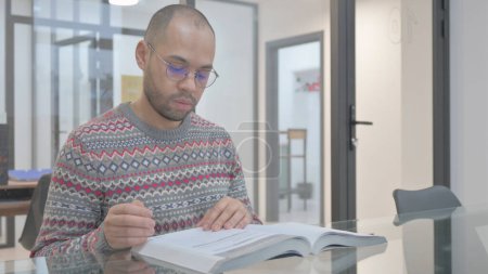 Téléchargez les photos : Jeune homme hispanique lisant le livre au bureau - en image libre de droit
