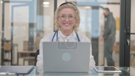 Téléchargez les photos : Senior Femme médecin sourire à la caméra tout en travaillant sur ordinateur portable - en image libre de droit