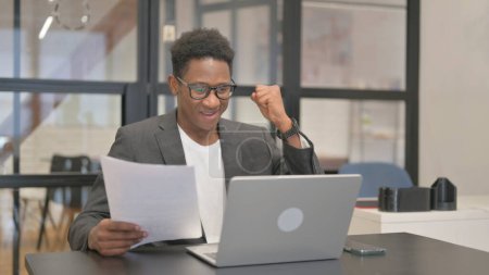 Téléchargez les photos : Afro-Américain Célébration tout en travaillant ordinateur portable et documents - en image libre de droit