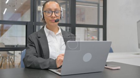 Téléchargez les photos : Hispanique femme d'affaires avec casque regardant caméra tout en travaillant sur ordinateur portable - en image libre de droit