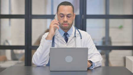 Téléchargez les photos : Médecin de course mixte Parler au téléphone tout en travaillant sur ordinateur portable - en image libre de droit