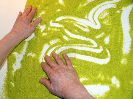 Foto de Manos de una anciana sobre una mesa blanca con arena verde, terapia de arena para habilidades motoras finas de los dedos, rehabilitación después de un derrame cerebral - Imagen libre de derechos