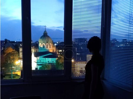 silueta de una chica en la ventana de un cuarto oscuro mirando a la ciudad de la tarde.