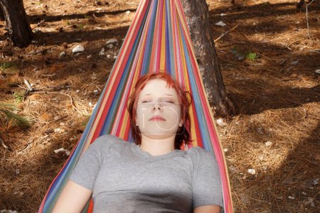 red-haired young woman lying eyes closed in a colorful hammock outdoors.