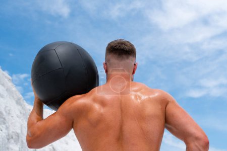 Téléchargez les photos : Un homme très fort tient sur ses épaules une grosse boule noire pour s'entraîner. La vue de l'arrière - en image libre de droit