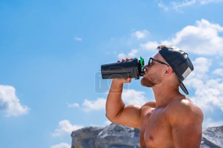 Téléchargez les photos : Un athlète masculin dans une casquette de baseball boit une boisson énergisante dans une bouteille contre un ciel bleu avec des nuages - en image libre de droit