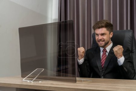Photo for A joyous and successful businessman celebrates with a beaming smile and clenched hands while looking at the monitor. Embrace the essence of triumph and happiness! - Royalty Free Image