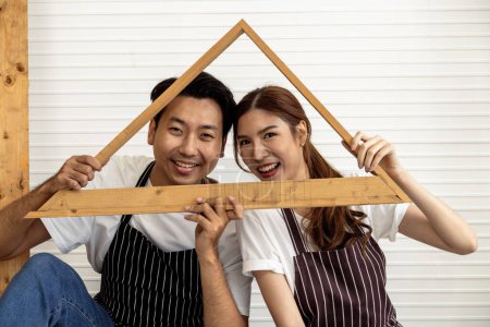 Photo for A couple in striped aprons and jeans holds a wooden house frame indoors. Their hands join together symbolizing unity and teamwork both in love and the journey of building their future home. - Royalty Free Image