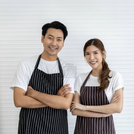 Photo for Two young adult Asians working as baristas. Dressed in white shirts and striped aprons. They are standing against a white wall background with arms crossed. - Royalty Free Image