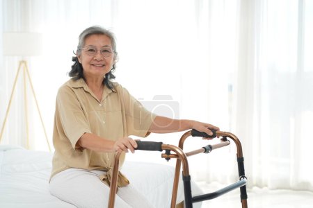 Portrait of Cheerful mature woman sitting on bed and mobility walker, looking at camera and smile with happy, copy space